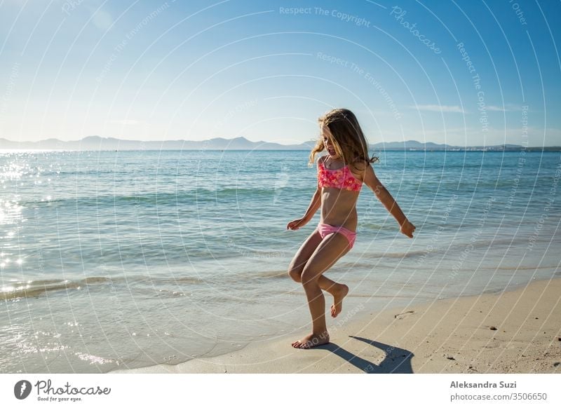 Cute happy little girl running along the beach in swimming suit, jumping over waves. Beautiful summer sunny day, blue sea, picturesque landscape. Majorca, Spain