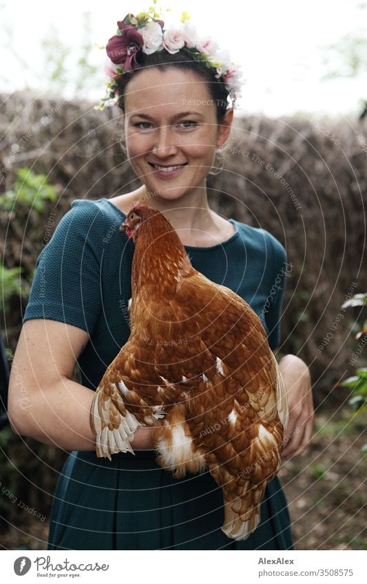 Young woman with a wreath of flowers in her hair stands in the chicken run holding a brown chicken in her arms Central perspective Shallow depth of field Day