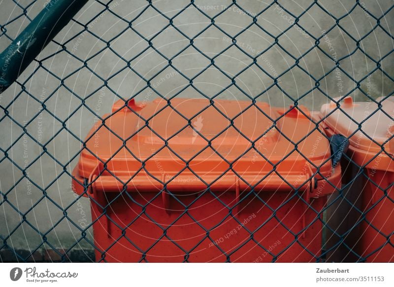 Garbage can in orange behind green wire mesh fence dustbin Orange Wire netting fence Trash Disposal city cleaning Fence little story lid Recycling Plastic