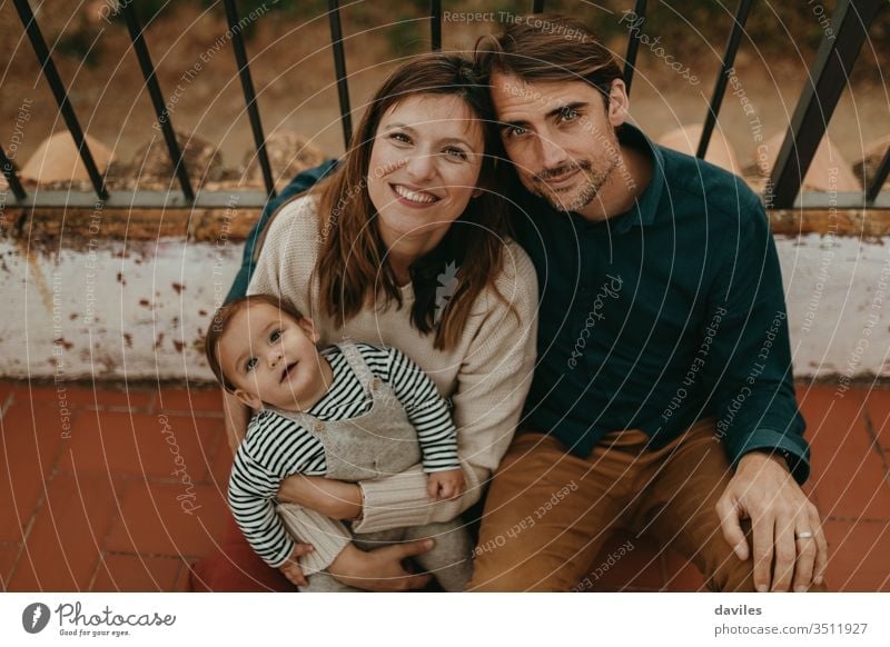 Lovely portrait of handsome man and beautiful woman with their baby son, looking at camera, while sitting on their home terrace floor. husband lovely cute