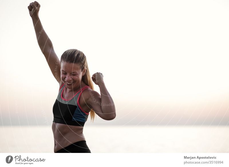 Young woman standing by the sea with arms raised in triumph achievement action active activity athlete beautiful challenge endurance evening exercise female fit