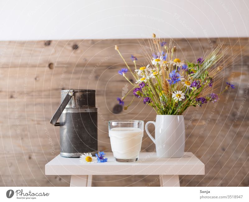 Ein Glas frische Milch, ein Strauß Wiesenblumen in einer Vase und eine alte Milchkanne auf einem weißen Hocker. Ländliches Motiv Trinkglas Blumen Rustic Alt
