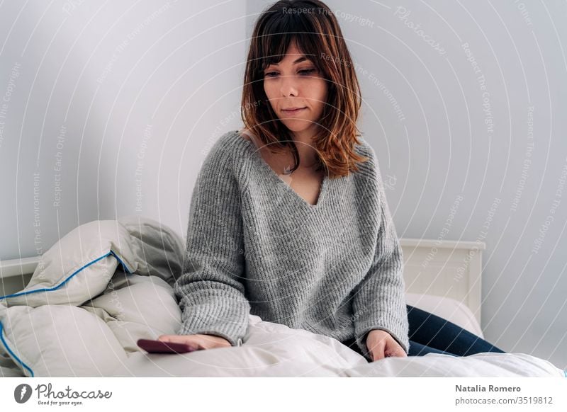 A young woman is sitting on the bed. She is holding her phone and looking at something on it. She looks worried. Concept stay home. quarantine internet pandemic