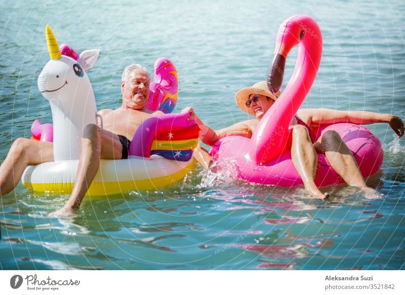 Elderly couple having fun on inflatable flamingo and unicorn. Funny active pensioners happy together enjoying summer vacation on the beach in Europe, laughing, playing the fool, splashing water.