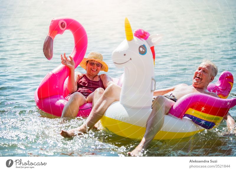 Elderly couple having fun on inflatable flamingo and unicorn. Funny active  pensioners happy together enjoying summer vacation on the beach in Europe,  laughing, playing the fool, splashing water. - a Royalty Free