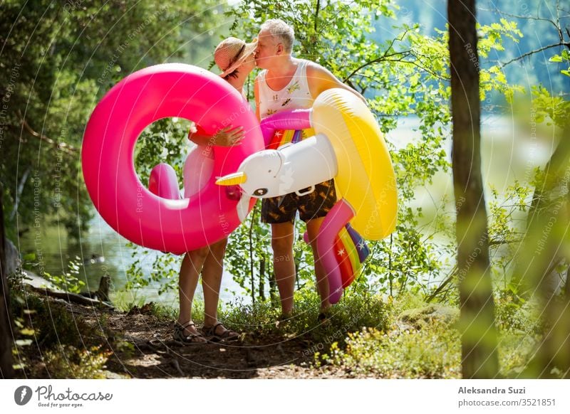 Elderly couple walking a forest path along the seashore holding giant inflatable flamingo and unicorn. Funny active pensioners enjoying summer vacation on the beach in Northern Europe