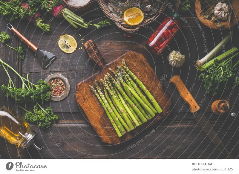 Fresh organic asparagus on wooden cutting board. Copped herbs, olive oil, lemon and seasonings. Preserved hot peppers in glass jar on dark rustic background, top view. Healthy lifestyle. Home cooking