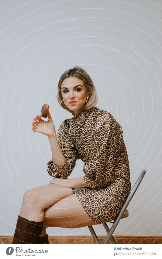 Cheerful stylish woman eating donuts smile style model sit chair bite female fashion trendy dress leopard print doughnut dessert pastry tasty chocolate food