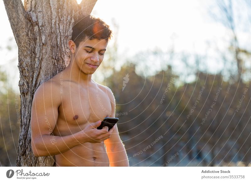Cheerful ethnic sportsman using smartphone during break park tree sit smile shirtless male athlete happy hispanic trunk browsing relax healthy muscular guy fit