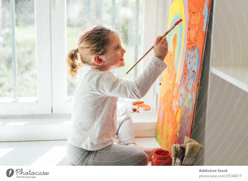 Little girl painting picture with paintbrush canvas draw rainbow creative colorful orange sill casual window art hobby imagination bright sit indoors