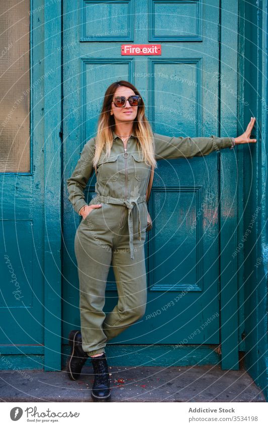 Cheerful woman in sunglasses standing near fire exit door trendy doorway style cheerful turquoise colorful bright overall hand in pocket boot positive