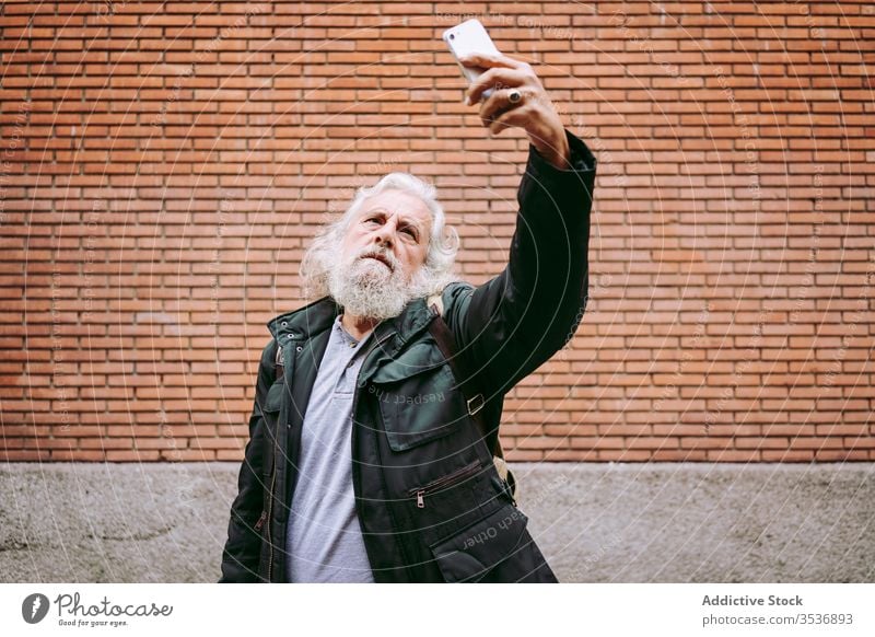 Senior male hipster taking selfie near brick wall in city senior man smartphone serious street using elderly aged gray hair long hair masculine handsome