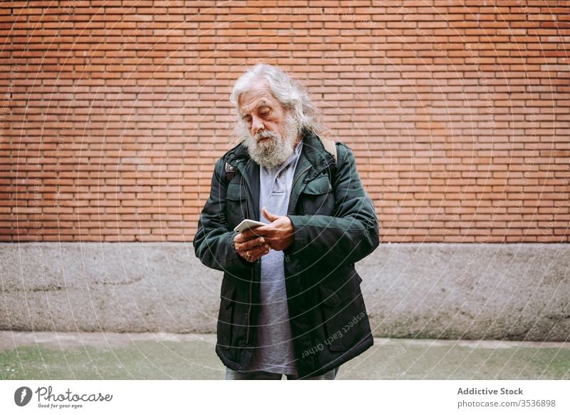 Male hipster using mobile phone near brick wall senior man smartphone city serious street male elderly aged gray hair long hair masculine handsome camera casual