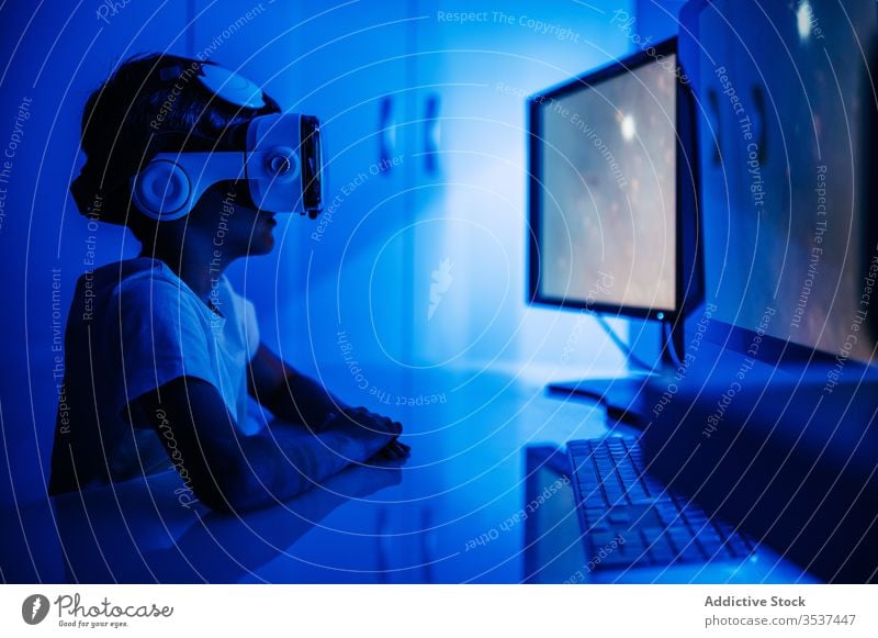 Unrecognizable boy in VR headset sitting in front of computer vr desktop monitor entertainment video using device gadget glasses wireless watch screen