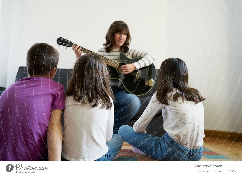 Cheerful woman playing guitar for kids in living room at home group sofa cheerful acoustic floor carpet together leisure relax sit music fun comfort hobby smile