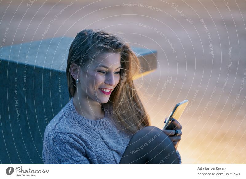 Smiling female browsing smartphone while resting in park at sunset woman happy cheerful glad using watching social media stone bench recline smile internet