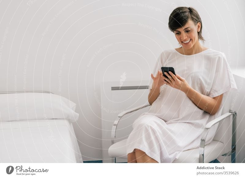Happy patient using smartphone in hospital ward woman smile chair sit bed female modern robe cheerful white browsing clinic device gadget internet relax rest