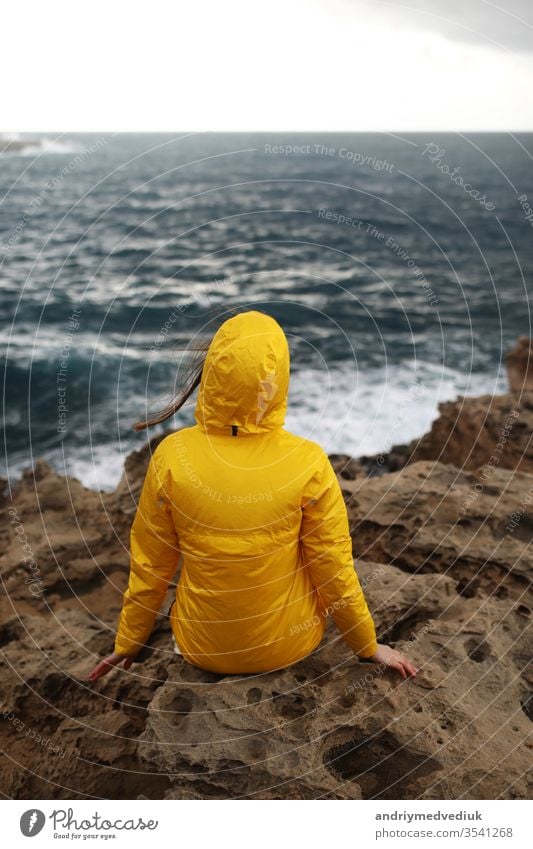 young woman dressed in yellow raincoat sitting on the cliff looking on big waves of the sea while enjoying beautiful sea landscape in rainy day on the rock beach in cloudy spring weather