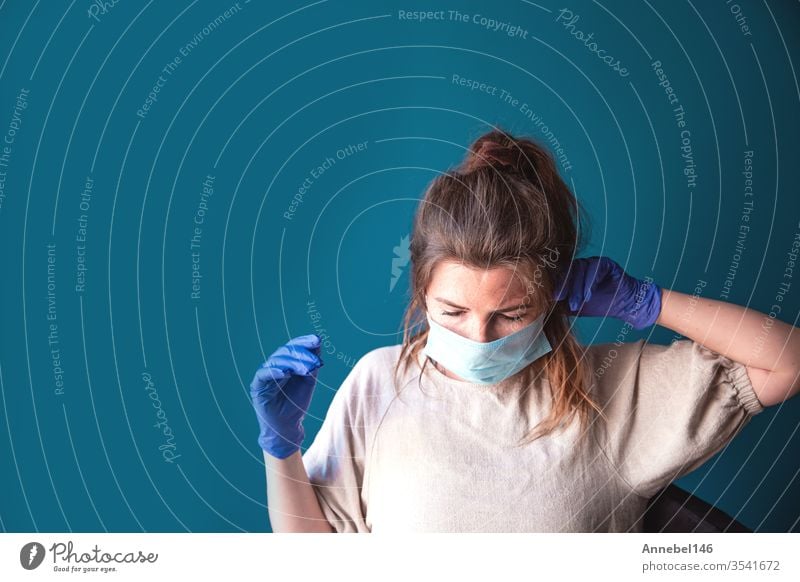 Young woman wearing protective gloves and face mask inside a home in quarantine looking bored and sad, for Covid-19 Coronavirus, with blue background medical