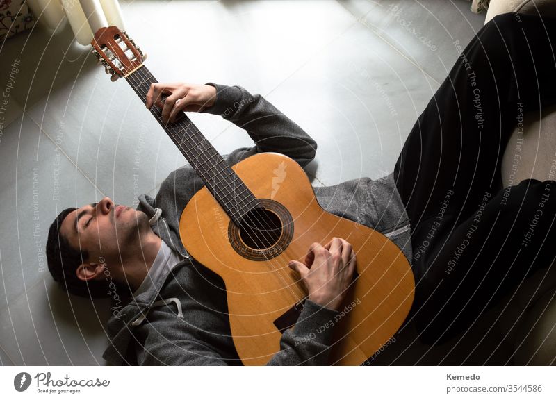 Relaxed man lying on the floor playing guitar near a bright window at home. Guitarist enjoying the music, harmonious moment. melancholy gloomy quarantine relax