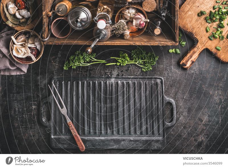 Grill or BBQ food background with empty cast iron grill griddle and meat fork  on rustic kitchen table. Wooden box with seasonings. Kitchen utensils.Top view. Place for design