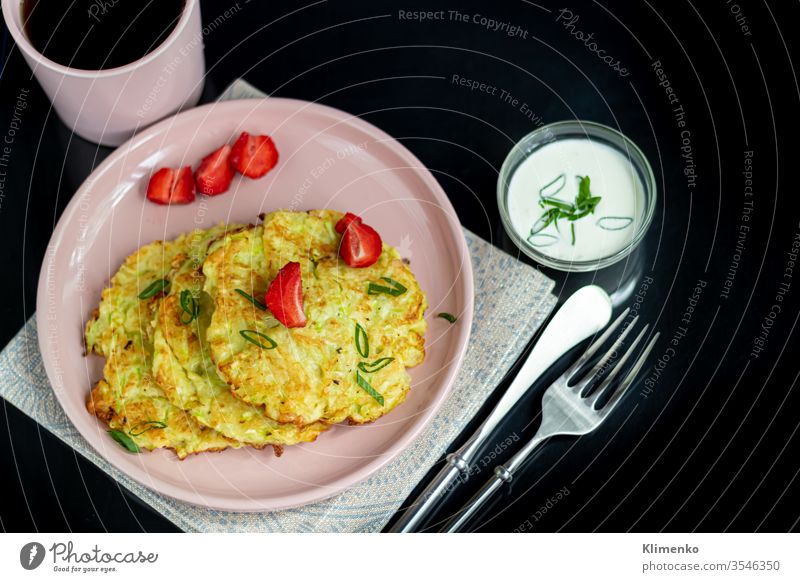 Zucchini fritters, vegetarian zucchini fritters, served with fresh herbs and sour cream. Garnished with strawberries and green onions. On a dark background.