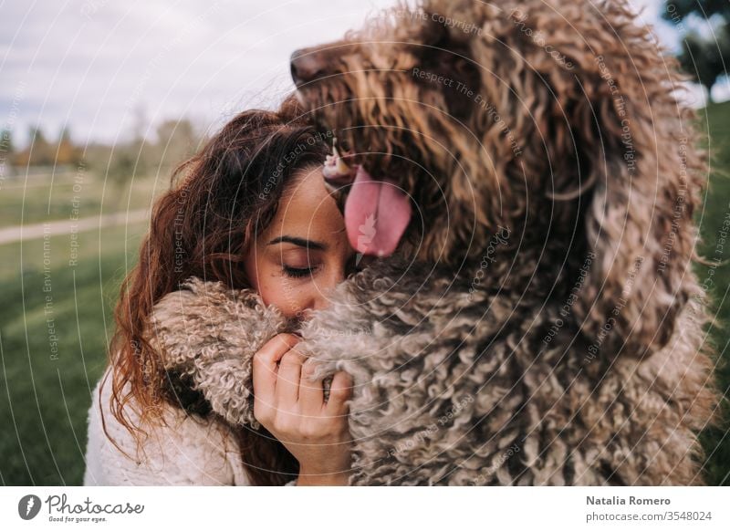 A beautiful woman is in the meadow with her dog. They are hugging. She has her eyes closed while the doggy is looking far. They are enjoying their company. The pet is a Spanish water dog.