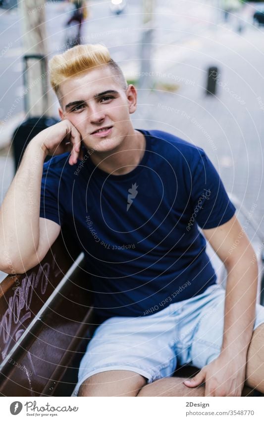 Portrait of a young teenage male with blonde bangs sitting on a bench in the street while looking at the camera person outdoor teenager man lifestyle caucasian