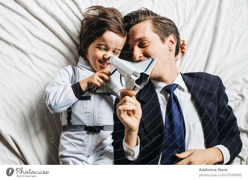 Content ethnic son and father lying on bed together astronaut costume boy dad play toy having fun diverse multiracial spacesuit parent spaceship smile kid
