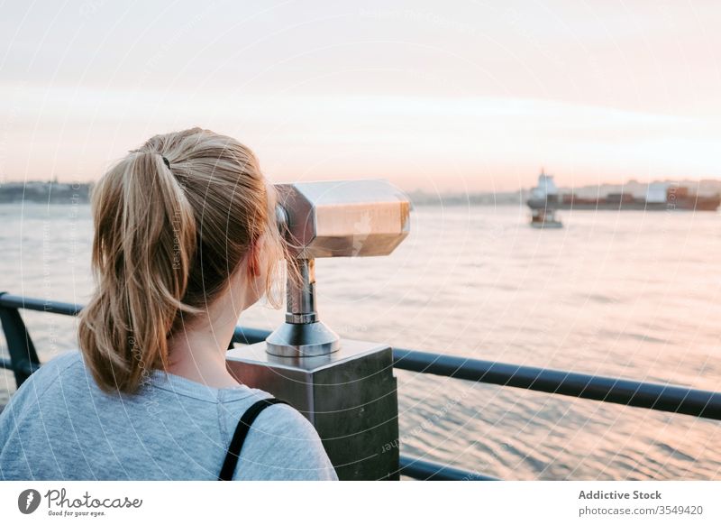 Young woman admiring view with binoculars platform observe sunset reflection magnificent sea promenade pink light calm horizontal traveler shore nature scenic