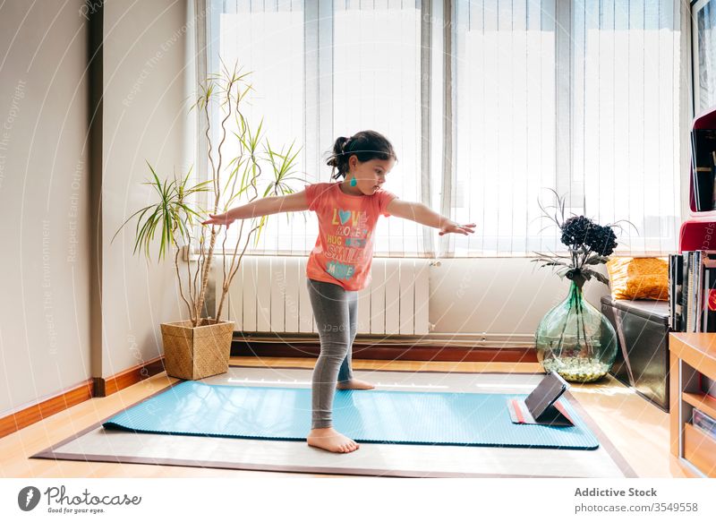 Joyful little girl doing yoga in light room kid pose mat video tablet tutorial balance practice at home stress relief watch harmony stretch flexible spirit