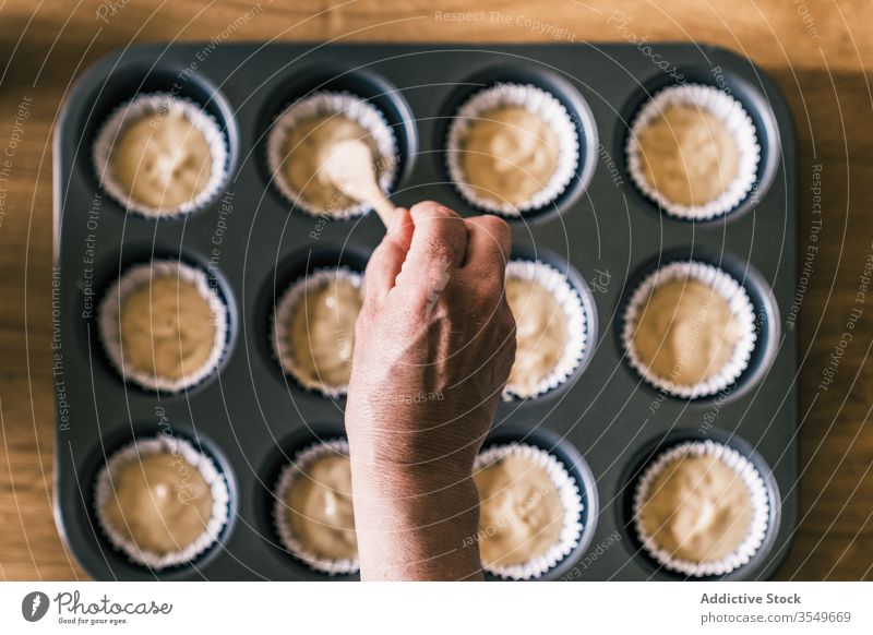 Crop woman filling muffin cases with dough tray housewife pour batter elderly spoon female cupcake liner kitchen senior home confectioner cook prepare paper