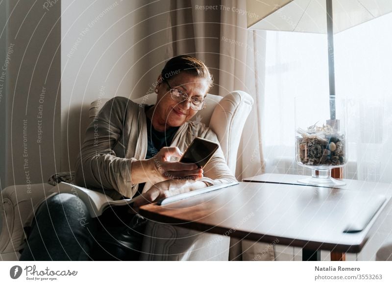 An older woman is sitting on the sofa with a book. She is typing on her phone while smiling. She is protecting herself and staying home. Pandemic concept.