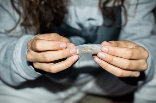 Young woman in tracksuit rolling a marijuana joint in the street at night. Details of hands rolling cannabis cigarette. girl young person teen smoke detail