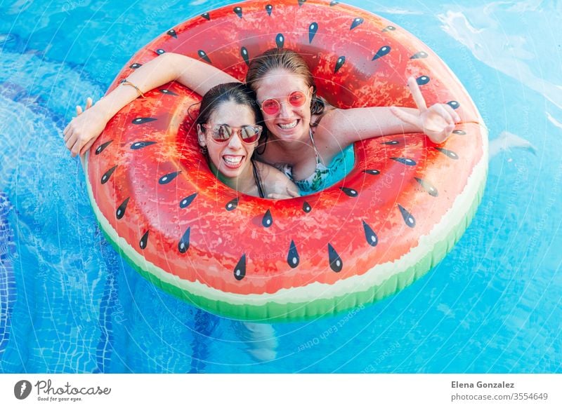 Two friends with sunglasses in inflatable watermelon ring. Young women enjoy the summer at swimming pool. Hand victory sign gesture. Fun time ladies females