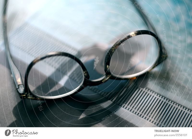 Viewed closely, it is a pair of glasses lying on a magazine, the title of which shows a mermaid with a bathing cap sitting in the sun at the edge of the pool and casting a strong shadow