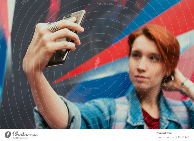 Young adult woman with dyed red hair doing selfie with her smartphone, selective focus, toned image happy fashion social media hand wall graffiti beauty concept