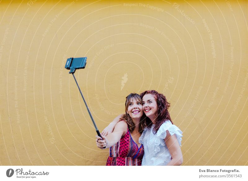 two friends or sisters taking a picture outdoors with mobile phone over yellow background. technology and lifestyle concept selfie city urban fun stick smiling
