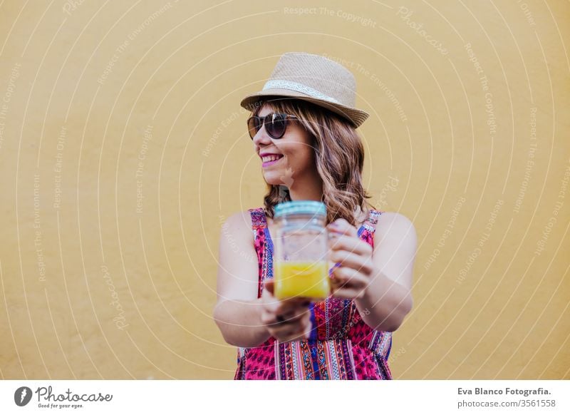 portrait of young caucasian woman outdoors over yellow background enjoying a cup of healthy orange juice. summer time city lifestyle curvy real people real life