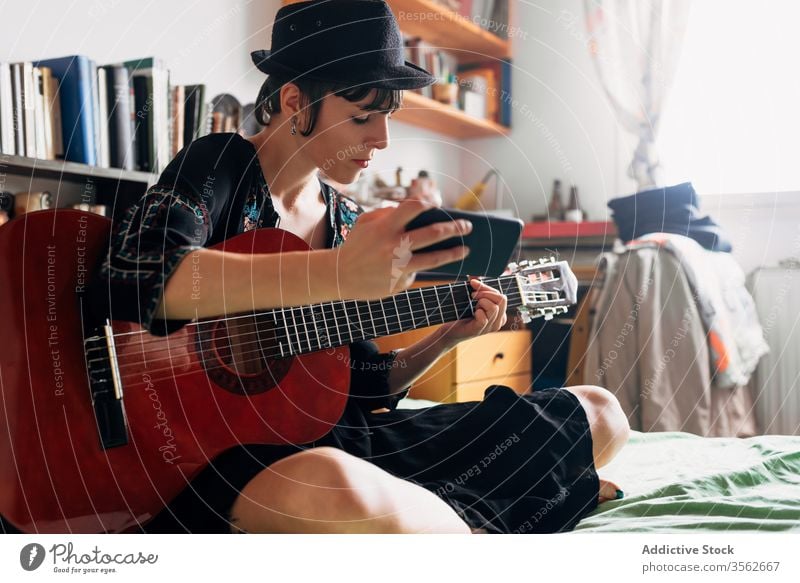 Woman with guitar and smartphone at home woman browsing acoustic trendy message using cellphone music female weekend style wear hat bed sit relax gadget device