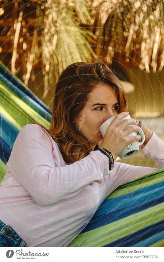 Relaxed woman with cup of coffee in hammock relax hot drink calm drinking barefoot courtyard enjoy female summer holiday vacation weekend mug casual outfit