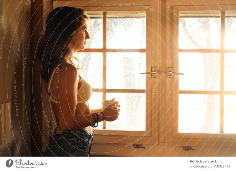 Relaxed woman with coffee near window in morning relax enjoy weekend cabin serene beverage female jeans bra cup hot drink stand rest young cottage wooden lady
