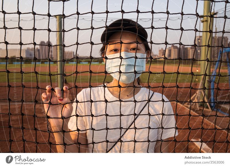 Young woman in protective mask standing behind net fence on sports ground coronavirus restriction prevent covid young sporty female asian ethnic safety