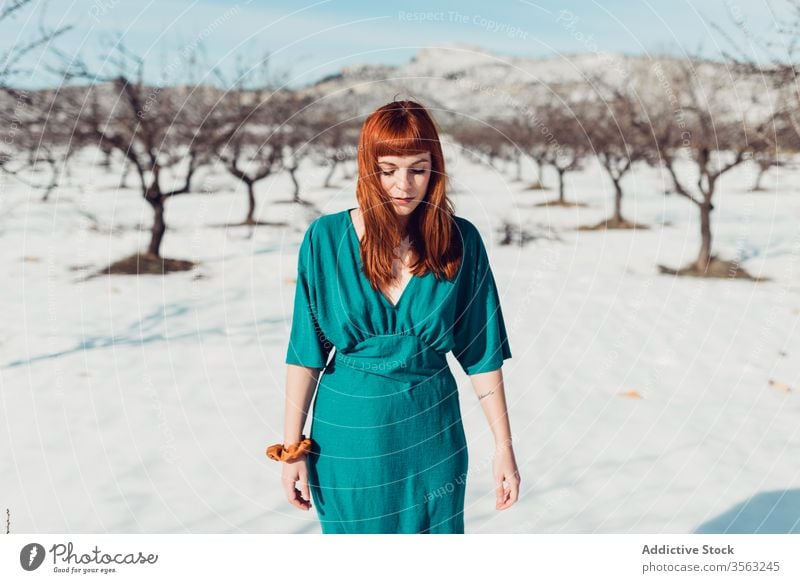 Young beautiful female with long red hair in turquoise outfit looking down while standing in snowy field in sunny day woman style trendy sensual redhead young