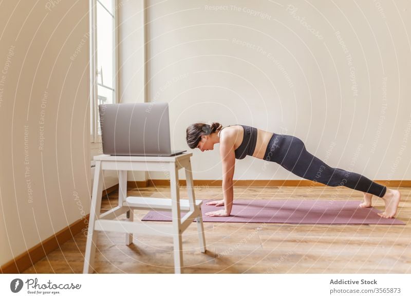 Fit female standing in plank pose at home woman yoga practice wellness healthy lifestyle fitness laptop mat using device gadget pc asana kumbhakasana sportswear