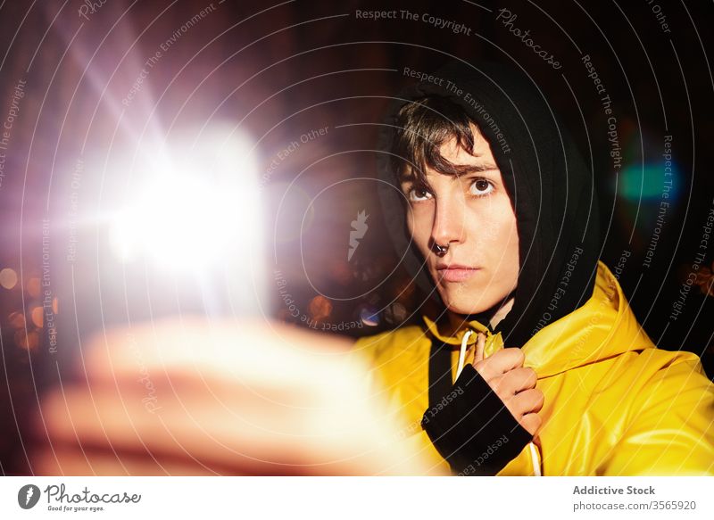 Young serious woman in hood photographing with smartphone at dark night light ray photography use raincoat take shot tranquil appearance travel calm search