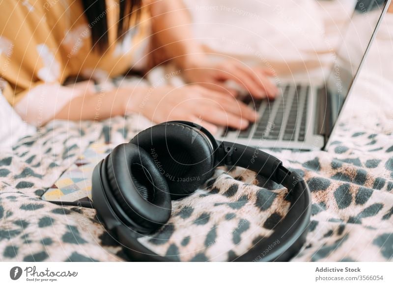 Crop woman browsing laptop with headphones smartphone bed chill home weekend comfort using typing keyboard lying lifestyle relax rest cozy social media bedroom