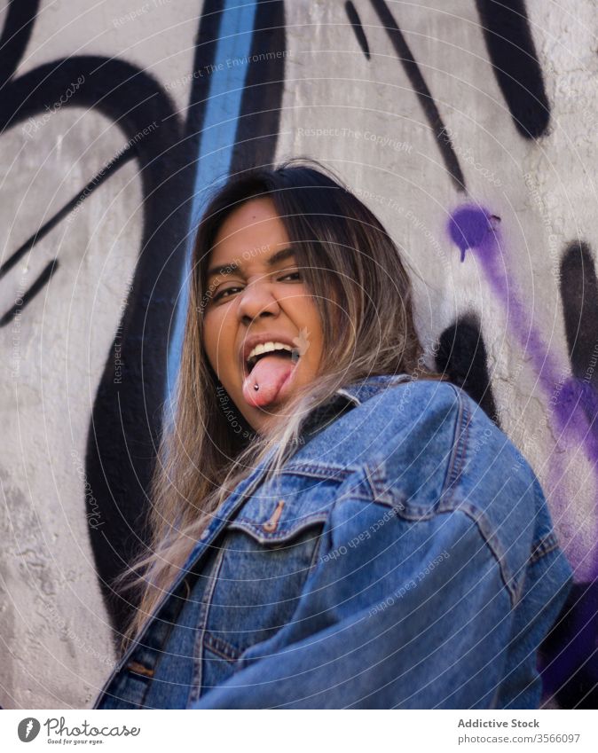 Naughty ethnic female millennial with tongue out on street woman rebel naughty piercing graffiti wall hipster denim jacket cool trendy style outfit weekend