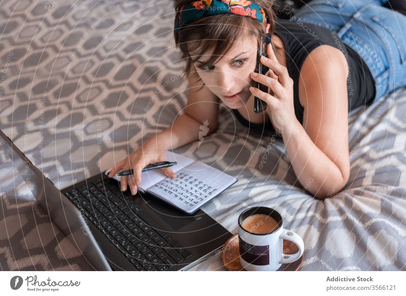 Young female freelancer talking on smartphone on bed woman self employed speak notebook laptop using young entrepreneur student learn education bedroom work job