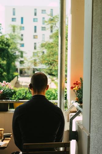 Man in black clothes with headset in his ear sits on the balcony in the sun and looks outside Balcony Sun good weather sunshine Rear view planted flowers Sit